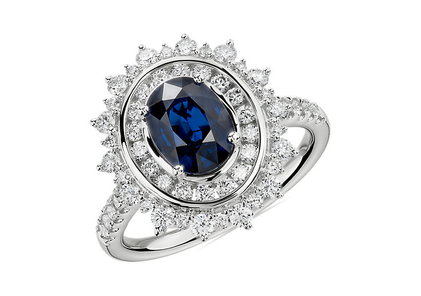 Un anillo de compromiso con zafiro ovalado decorado por un halo doble de diamantes en un diseño en forma de rayos engarzado en oro blanco