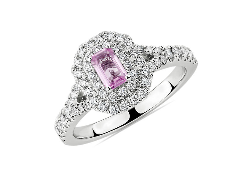 Un anillo de compromiso de estilo romántico con un zafiro rosado de talla esmeralda en el centro rodeado por un halo doble de diamantes y diamantes incrustados en la banda de oro blanco