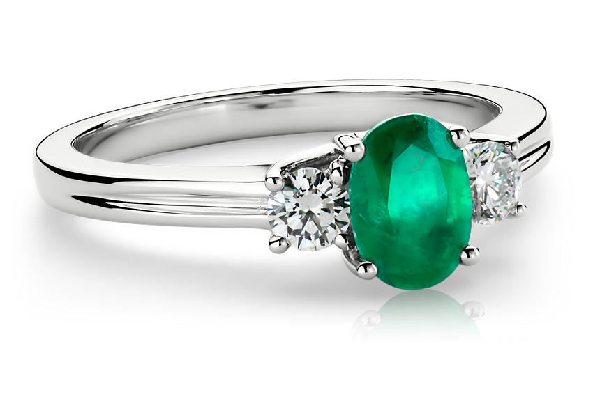 An oval cut emerald center is framed by two round diamonds in a modern engagement ring with clean lines and soft curve detail of the white gold setting