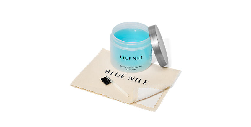 Un set de limpieza de joyas con una botella abierta de limpiador líquido azul, un paño de color crema con el logotipo de Blue Nile en azul marino, y un cepillo suave de color negro con mango de acrílico transparente.
