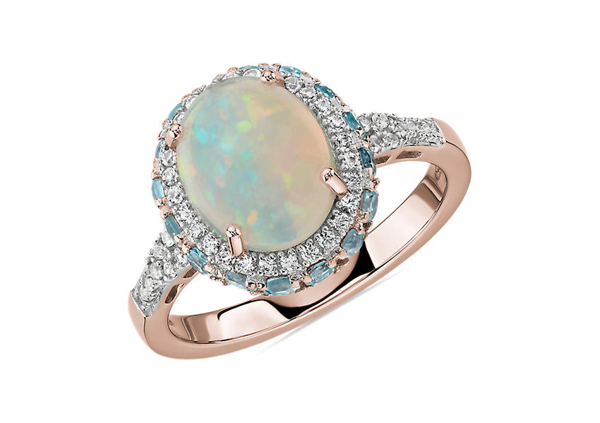 A cabochon cut oval opal engagement ring framed by a double halo of blue topaz and white sapphires set in rose gold