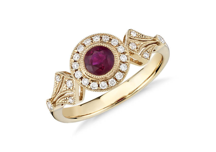 Un anillo de compromiso con rubí redondo de inspiración retro decorado con un halo milgrain con diamantes en pavé a lo largo de un engarce de oro amarillo