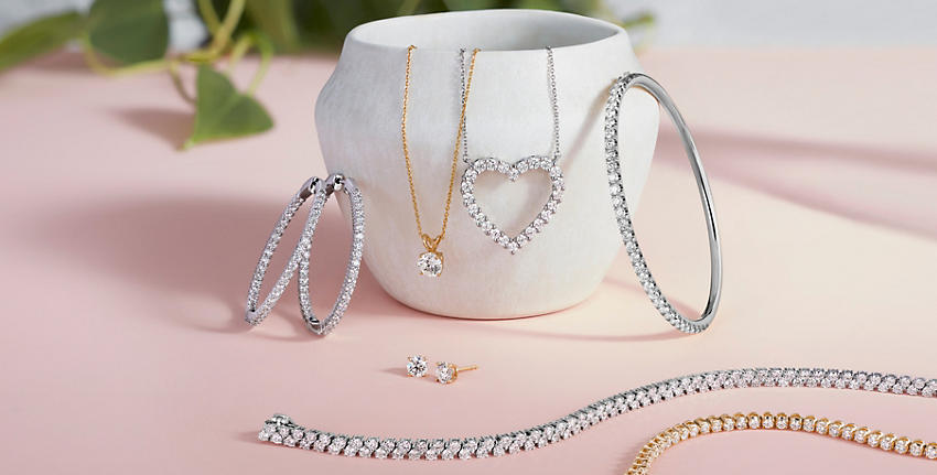 White vase on pink surface with diamond hoops, diamond pendant necklace, diamond heart necklace, diamond bangle, diamond stud earrings, and diamond tennis bracelets.
