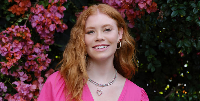 Pale female with red hair wearing magenta v-neck top and garnet studs, diamond hoops, heart necklace, and two eternity necklaces.