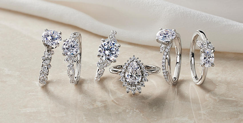 Seis diseños de anillos de compromiso con diamantes: anillo de compromiso asimétrico con motivo floral romántico y diamantes, anillo de compromiso romántico de diseño torcido y diamante en forma de pera, anillo de compromiso romántico de diseño torcido con diamantes redondos y en forma de pera, anillo de compromiso con motivo floral y diamantes redondos y de talla marquesa, anillo de compromiso asimétrico con diamantes de talla redonda y marquesa y anillo de compromiso con halo ballerina de diamantes redondos, en forma de pera y talla marquesa sobre una superficie de color beige.