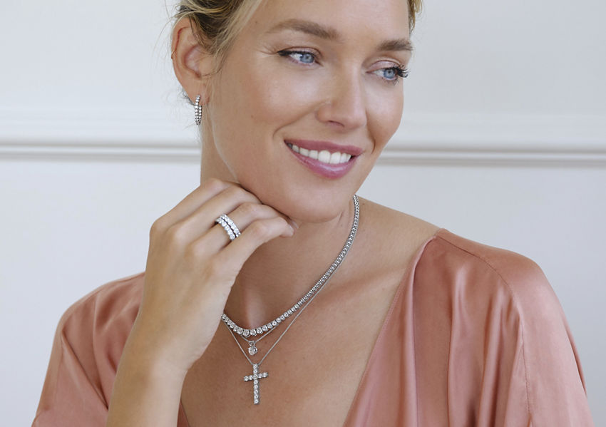 Woman with blond hair wearing satin dusty rose blouse, diamond hoops, eternity necklaces, bracelets, and rings. Desktop version also shows woman's wrists wearing diamond eternity bracelets.