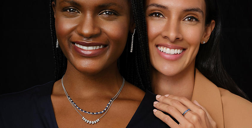 Two females with black hair and brown eyes smiling and facing camera. Left female wears Diamond Line Drop Earrings; Diamond Pear Shape Smile Necklace in 14k White Gold; Oval Sapphire and Round Diamond Necklace; and black V-neck top. Right female wears Channel Set Blue Sapphire Ring; Diamond Stud Earrings; and beige blazer.