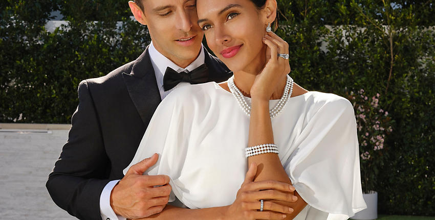A male and female dressed in formal wear smile and face camera. Female, at right, wears Classic Six-Prong Solitaire Engagement Ring; Pointed Diamond Insert; French Pavé Diamond Ring; Cushion-Cut Diamond Double Halo Drop Earrings; Triple-Strand Freshwater Cultured Pearl Strand Necklace; Triple-Strand Freshwater Cultured Pearl Bracelet; and white dress.